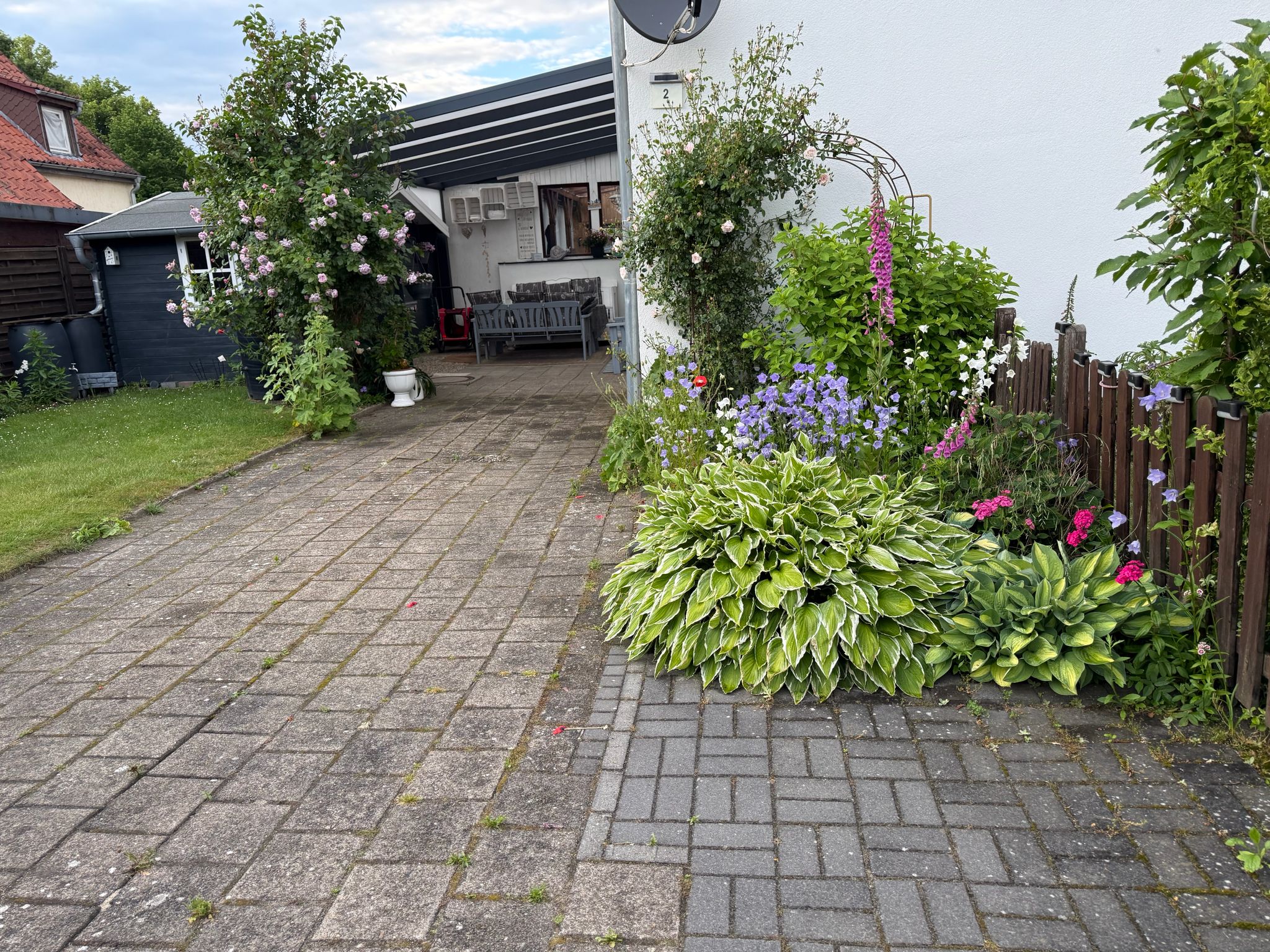 Eingezäunter Garten mit Rosenbogen, überdachter Terrasse und blühendem Staudenbeet